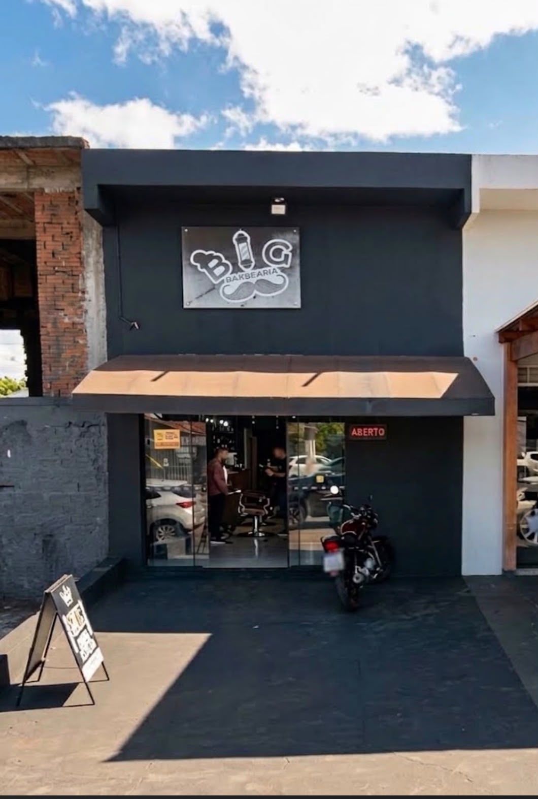 Fachada preta da Big Barbearia com toldo marrom, porta de vidro e a placa de aberto acesa